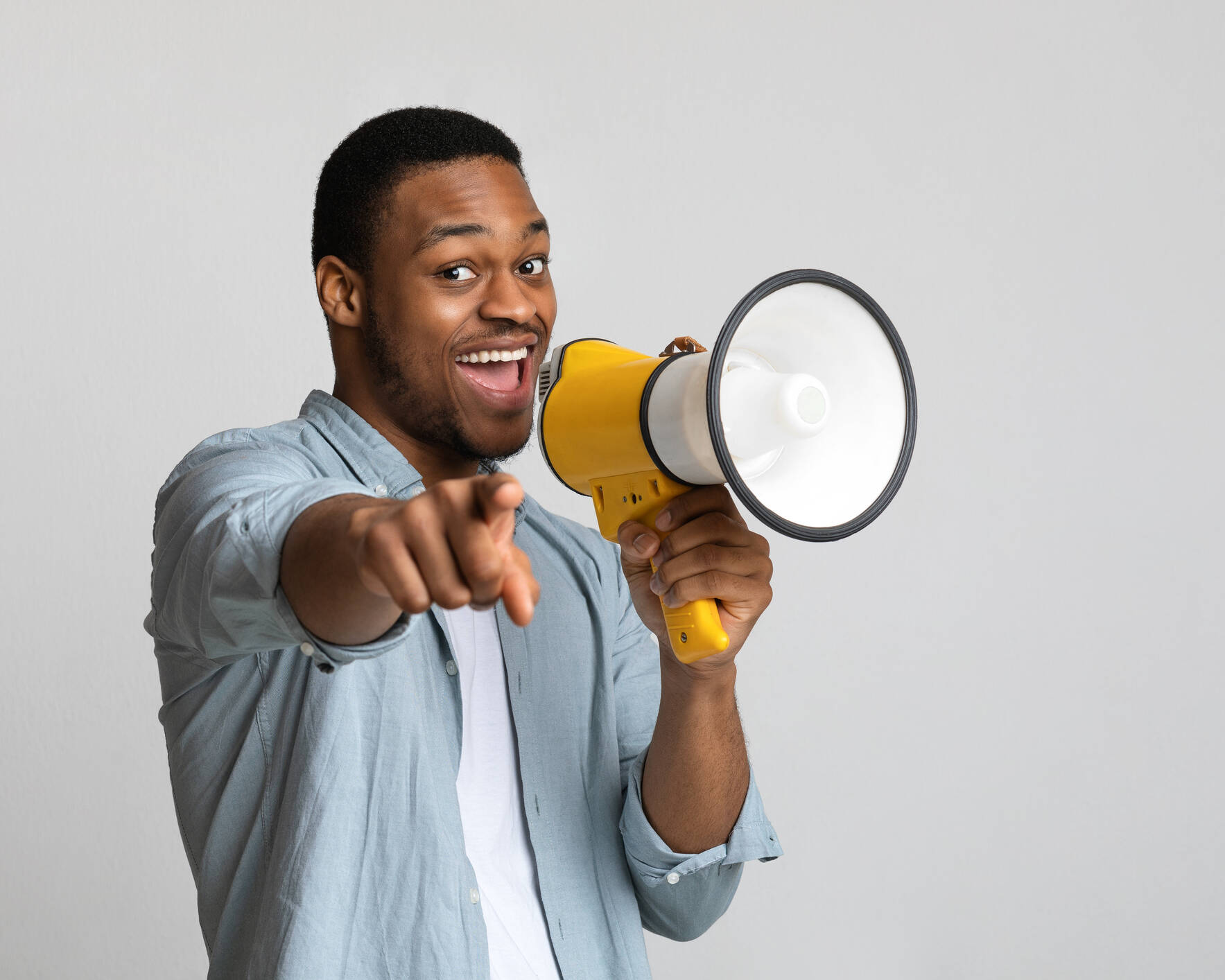 An image of a Positive looking African American guy shouting in megaphone and pointing at camera over grey background, copy space. Happy black man screaming with loudspeaker, cheering up, making advertisement